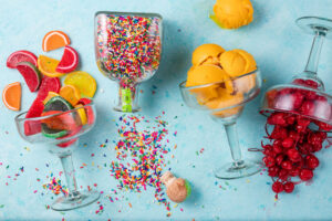 Colorful candy slices in a cocktail glass, sprinkles in another, with scoops of yellow ice cream and fresh fruit on a blue surface.