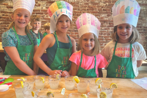 A group of children wearing green Sticky Fingers Cooking® aprons and colorful paper chef hats, standing behind a table with cups of lemonade.