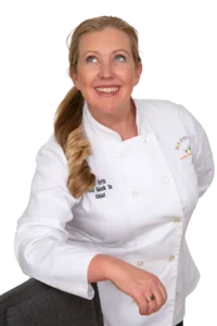 Erin Fletter, founder of Sticky Fingers Cooking®, smiling and looking upwards while wearing a white chef's coat.