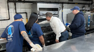 Four team members clean a commercial kitchen. One person points at the stove while others watch attentively.