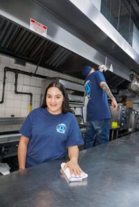 A woman in a navy uniform wipes a countertop, smiling. In the background, a man cleans an overhead kitchen vent.