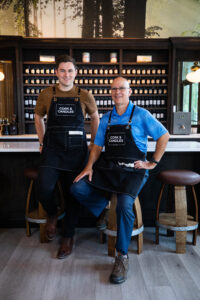 Kenny and Dave Straub, founders of Cork and Candles, wearing aprons and standing inside their candle bar.