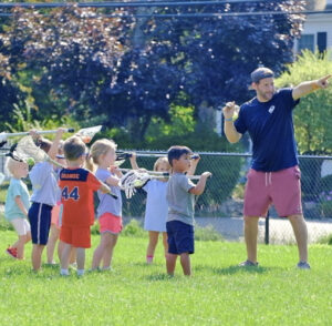 Coach instructs a group of young kids holding lacrosse sticks during an outdoor practice.